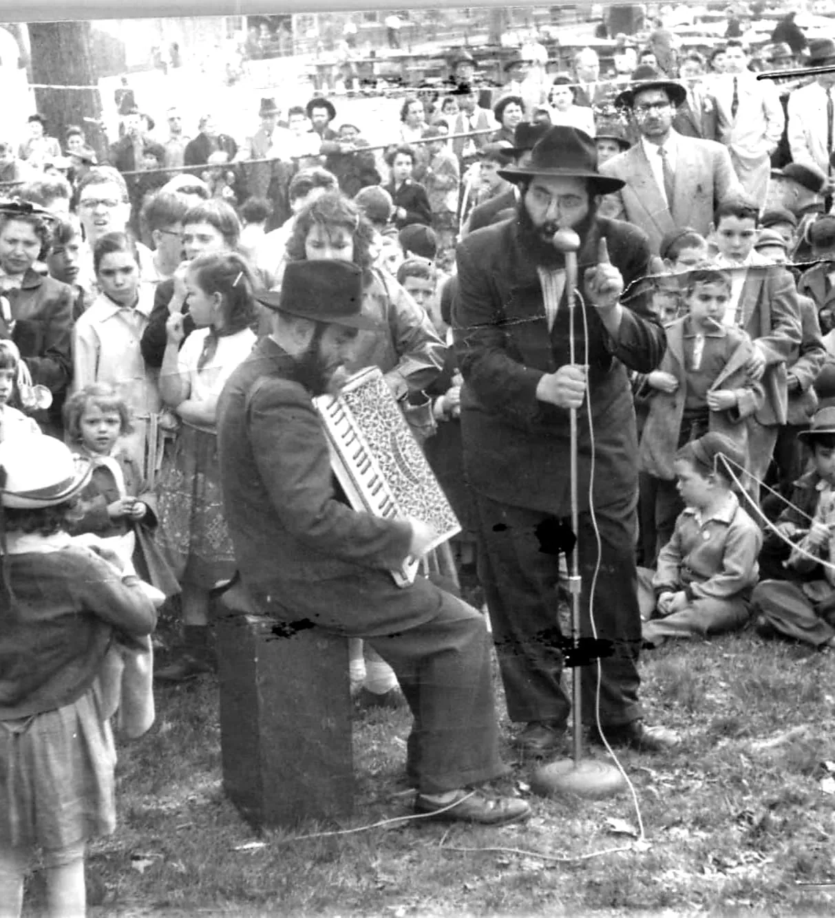 A crowd gathered outdoors around a speaker at a microphone and an accordion player